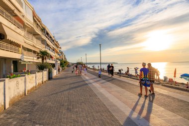 Bir grup insan okyanus kıyısında bir kaldırımdan aşağı yürüyor. Gökyüzü bulutlu ve güneş batıyor. 8 Ağustos 2024 Perea Yunanistan