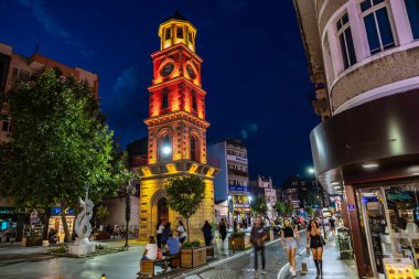 Gece çeşitli insanlarla dolu şehir meydanında aydınlatılmış saat kulesi. 2 Ağustos 2025, Canakkale Türkiye.