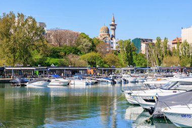 Güneşli bir günde cami ve yemyeşil yemyeşil manzaralı nehir kıyısına yanaşan tekneler. 3 Mayıs 2025 Romanya.