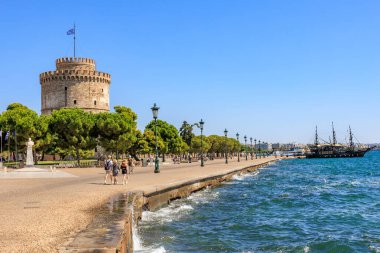 Bir grup insan büyük bir kule yakınında bir sahil boyunca yürür. Gökyüzü açık ve su sakin. 8 Ağustos 2024 Selanik Yunanistan