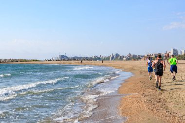 Arka planda şehrin silueti olan kumlu sahil boyunca koşan insanlar. 4 Mayıs 2025 Romanya.