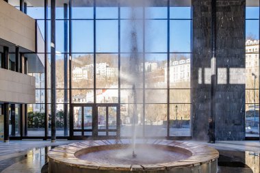 Camdan duvarı ve şehir manzarası olan görkemli bir çeşme. 18 Mart 2025, Karlovy Vary, Çek Cumhuriyeti.