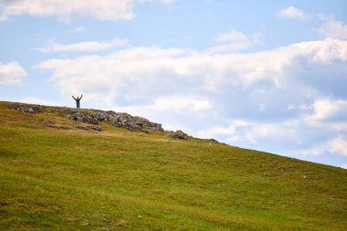 Bir kişi çimenli bir tepenin üzerinde duruyor. Kollarını kaldırır, gökyüzünü kucaklarlar. Bulutlar tepenin üstünde tembelce sürükleniyor. Yeşil yamaçlar yavaşça uzağa yuvarlanıyor.