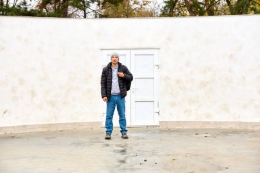 Adam beyaz duvarın önünde durup el kol hareketi yapıyor. Ceket, kot pantolon ve bere giyiyor. Arka planda sade kapı ve seyrek ağaçlar var. Ciddi ruh hali, sakin duruş, korunmasız ifade.