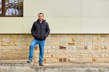 Adam taş duvara yaslanıyor. Koyu renk kabarık ceket ve kot pantolon giyiyor. Yumuşak, rahat ve cana yakın gülümsüyor. Doğal ışık, onun dostane davranışlarını vurguluyor. Yaşam tarzı ve kentsel yaşam için ideal