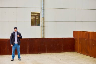 Adam endüstri binasının önünde duruyor. Sıradan bir ceket ve kot pantolon giyerek el hareketleriyle hareket ediyor. Yıpranmış çelik duvarlar, temiz beyaz panellerle tezat oluşturuyor. Metal boru pencerenin yanında dikey çalışıyor