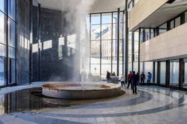 Modern bina avlusunda gayzer sergilenirken, ziyaretçiler buhar püskürmesini gözlemliyor. 18 Mart 2025, Karlovy Vary, Çek Cumhuriyeti.