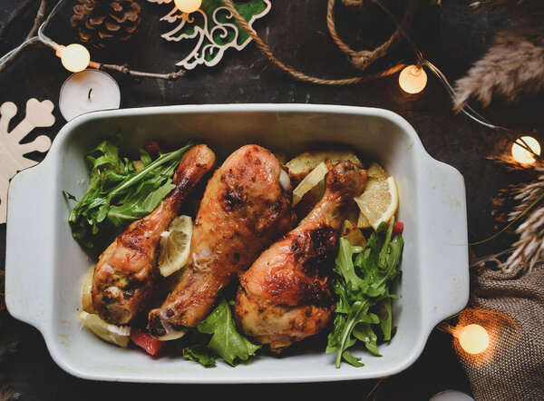 Winter festive food concept. Top view of dark vintage background, garland and candles. Christmas and New Year background. Baked chicken legs and potatoes.
