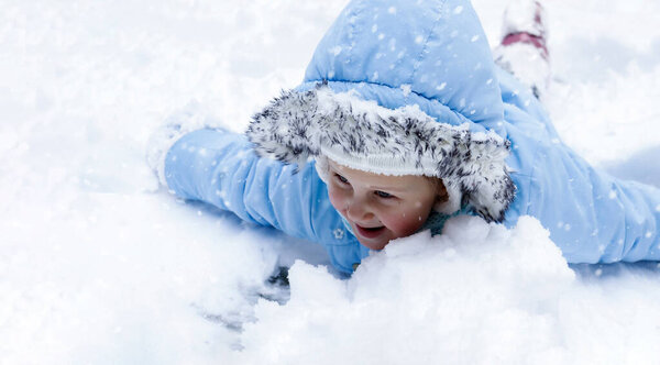 The baby is playing with the snow. Blue mittens and a blue jacket in the snow. The child lies and eats snow.