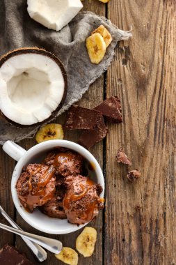 Çikolatalı hindistancevizli vejetaryen dondurmayı beyaz bardakta. Çikolatalı ve karamel kaplı dondurma topları. Ahşap arkaplan, kopyalama alanı.