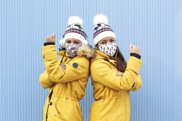 Two people wearing face masks and winter clothes isolated on a blue background. They are doing with their arms a gesture of force. Concept of the fight against the Covid-19 virus pandemic.