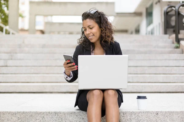 Young latina entrepreneur woman using a smart phone and laptop computer outdoors. Space for text.