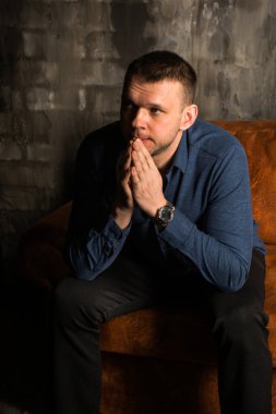 Mavi gömlekli düşünceli bir adam karanlık bir odada, eski bir kanepede, tuğla bir duvara karşı oturuyor..
