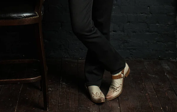 Gray cowboy boots on the feet of a man. The feet of a young man in black trousers and cowboy boots on an antique wooden floor.