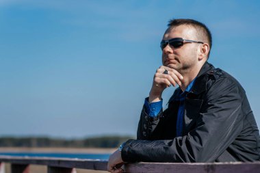 Siyah ceketli ve güneş gözlüklü genç bir adam eski bir ahşap köprünün üzerinde duruyor ve uzaklığa bakıyor. Nehir kıyısında genç, yakışıklı bir işadamı.