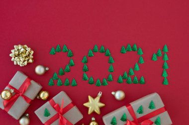 2021 yazıtları düz, küçük yeşil köknar ağaçları, kırmızı kurdeleyle yorgun hediye kutuları ve kırmızı arka planda yeni yıl süslemeleri. Yeni yıl kavramı