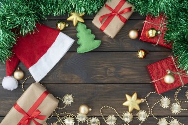 Noel ve yeni yıl süslemelerinin üst görüntüsü, Noel Baba şapkaları, hediye kutuları, altın toplar ve yıldızlar ve koyu ahşap arka planda yeşil süsler, merkezde kopyalanmış boşluk.