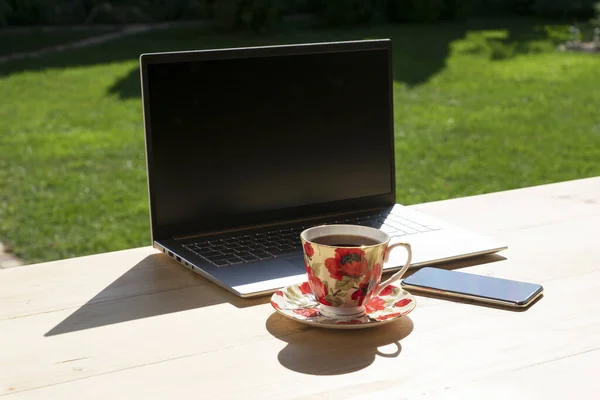 Laptop, telefon ve yeşil bahçedeki açık ahşap masada duran porselen çay standı. Serbest çalışmak ya da rahat bir yerde uzaktan öğrenmek. Uzak çalışma konsepti.