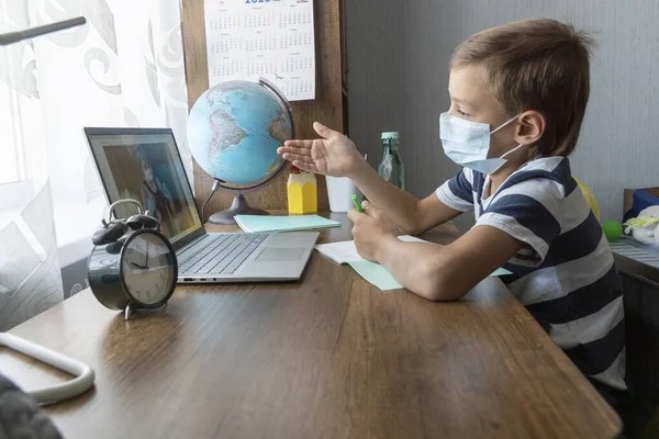 Tıp maskeli okul çocuğu evde dizüstü bilgisayar kullanarak online ders sırasında öğretmeniyle iletişim kuruyor. Uzaktan eğitim kavramı. Yeni gerçeklik.