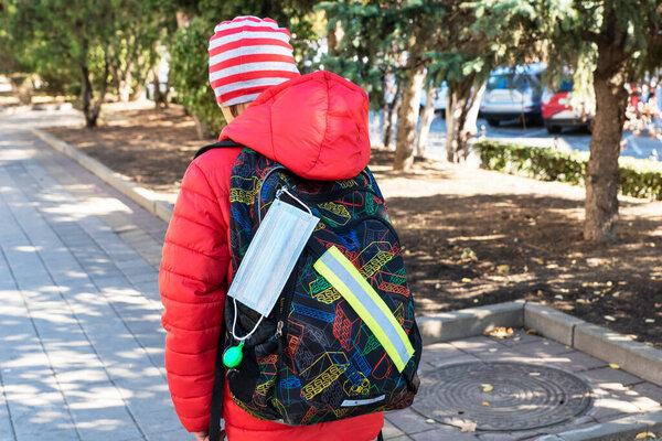 face medical mask is hanging on school backpack. Schoolboy is going to school throu park. Protective measures against viruses. Healthcare concept.