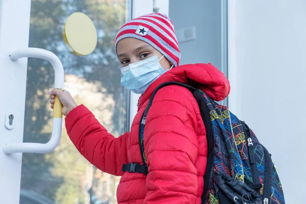 schoolboy in medical face mask looking into camera and pulling door before entering school. New reality, restrictions to prevent covid pandemic. Healthcare concept