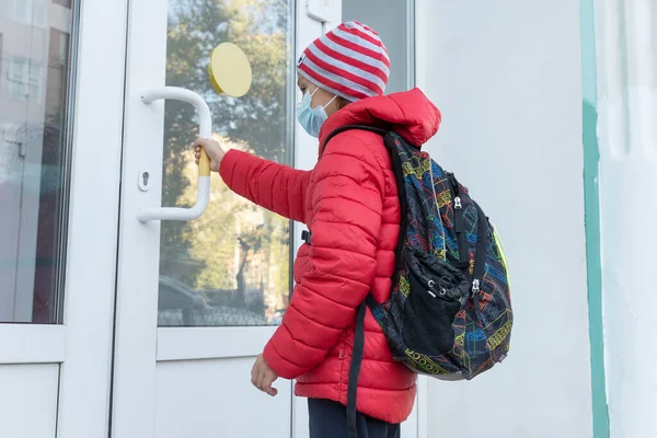 schoolboy in medical face mask is near door and pulls handle before entering school. New reality, restrictions to prevent covid pandemic. Healthcare concept