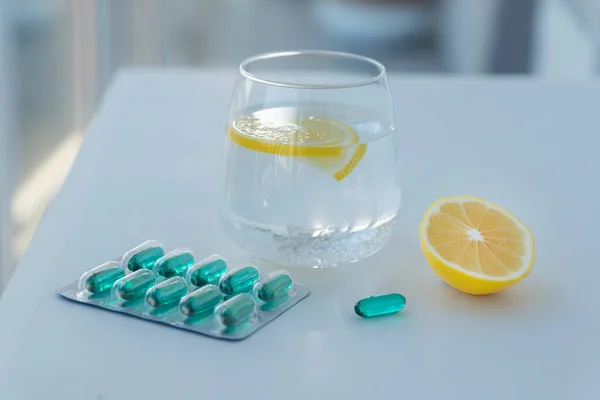 glass of water with two lemon slices, half of lemon, blister of blue medicine capsules on white table. Treatment and recovery measures. Healthcare concept.