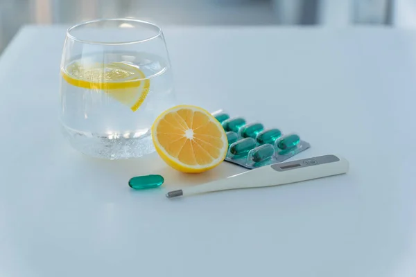 glass of water with lemon slices, half of lemon, blister of blue medicine capsules and termometer on white table in hospital. Treatment measures. Healthcare concept.