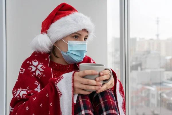 sad young female in face mask and santa hat covered with plaid hold cup of tea and look into window. Loneliness during holidays due to illness.