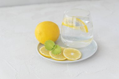Saf suyla şeffaf cam, limon dilimleri, nane yaprağı, arkaplanda beyaz tabakta duran bütün limon. Sağlıklı vitamin içeceği