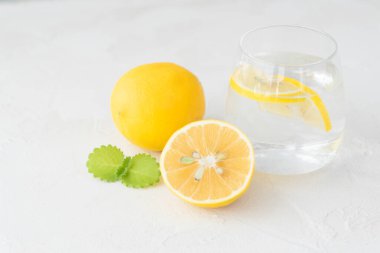 Saf suyla şeffaf cam, limon dilimleri, nane yaprağı, tam limon ve yarısı açık arkaplanda yatıyor. Sağlıklı vitamin içeceği