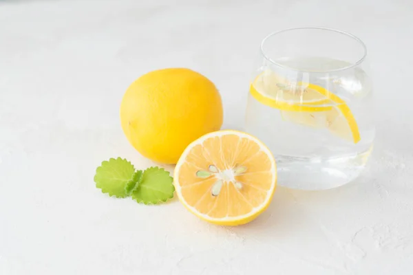 Saf suyla şeffaf cam, limon dilimleri, nane yaprağı, tam limon ve yarısı açık arkaplanda yatıyor. Sağlıklı vitamin içeceği