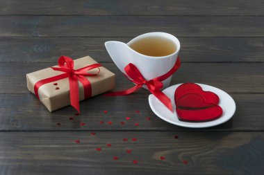 Paketlenmiş hediye kutusu açısı ve kırmızı kurdelelerle bezeli beyaz bir fincan çay, iki kalp küçük tabakta yatıyor. Sevgililer Günü konsepti