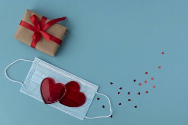 İki kırmızı kalp koruyucu tıbbi maske, paketlenmiş hediye kutusu ve küçük pırıl pırıl kalp konfetileri yakınlarda. Yeni gerçeklikte Sevgililer Günü