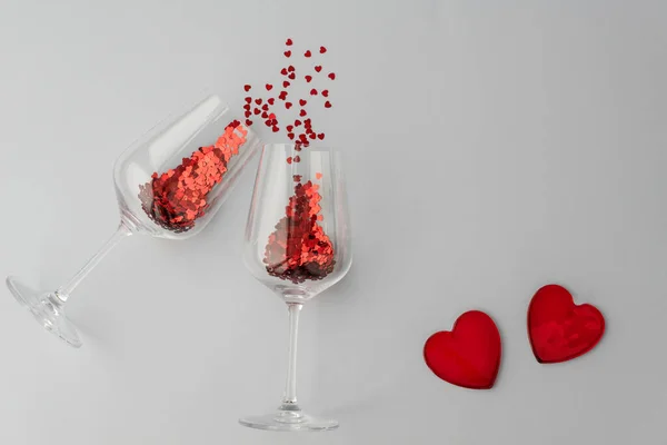 two wine glasses with little red heart sparkling confetti and two red transparent hearts in right corner on light background, lots of copy space for text. Love and valentines day concept