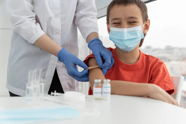 Tıbbi maske takan mutlu çocuk aşıdan sonra gözlerini kapadı. Doktor iğne yerine sıva, aşı, yeni gerçeklik aşısı yapıştırdı.