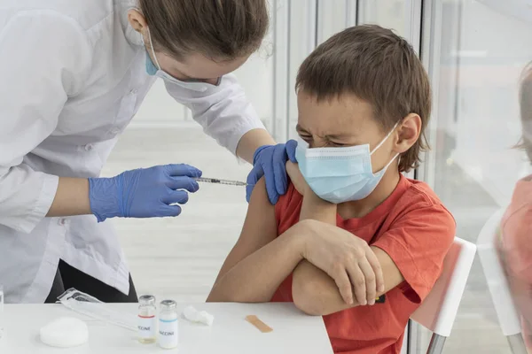boy in medical mask with closed eyes is afraid of injection. Female doctor is vaccinating young patient. Healthcare and vaccination concept.