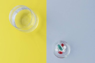 2021 'in arka planında farklı şekillerde ve renklerde haplar ve bir bardak suyla küçük yuvarlak tabakanın üst görüntüsü, gri ve sarı. Sağlık hizmetleri kavramı