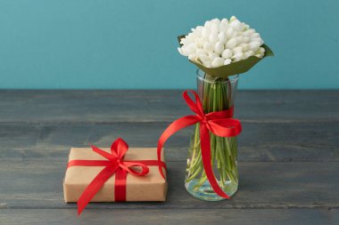 Küçük şeffaf vazodaki beyaz kar damlaları kırmızı kurdeleyle yorulmuş, paketlenmiş hediye kutusu açık mavi ve ahşap arka planda. Boşluk, selamlama kavramı