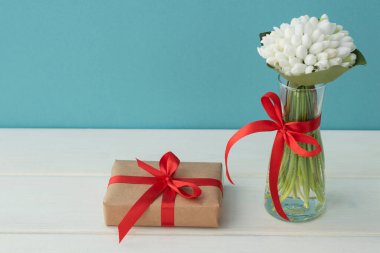 Küçük şeffaf vazodaki beyaz kardelen buketi kırmızı kurdeleyle bezeli hediye kutusu beyaz ahşap ve açık mavi arkaplanda. Boşluk, selamlama kavramı