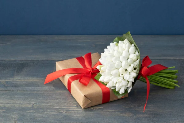 Beyaz bahar kardelenleri, koyu arkaplandaki paketlenmiş hediye kutusunda duran kırmızı kurdeleyle yoruldu. Bahar tatili, selam konsepti..