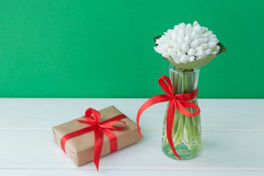 Küçük şeffaf vazodaki bahar çiçekleri kırmızı kurdeleyle bezeli yeşil ve beyaz ahşap arka planda el işi kağıt hediye kutusuna sarılı vazolar. Selamlar konsepti.