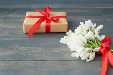 Kırmızı kurdeleli beyaz kardan küçük bir buket, koyu ahşap arka planda paketlenmiş hediye kutusu. Uzayı metin için kopyala. Bahar tatili, selam konsepti.. 