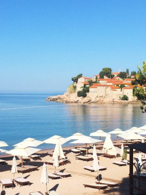 Karadağ 'ın Sveti Stefan kentinde güneş panjurları ve şemsiyeli plaj, ortaçağ şatosu, güzel mavi deniz ve parlak gökyüzü manzaralı. Güzel yerler konseptiyle seyahat etmek.