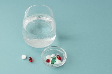 Büyük yuvarlak su bardağının, küçük şeffaf kapsülün, hapların ve kapsüllerin olduğu bir tabağın, kırmızı kapsülün yakınında yattığı bir beyaz hapın, sağlık ve iyileşme konseptinin yakın plan resmini çek.