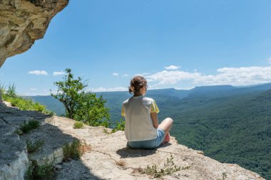 Young female sit on cliff on peak in high mountains looking over green mountains with feeling of relexation. Healthy lifestyle, adventure during hiking in mountains.