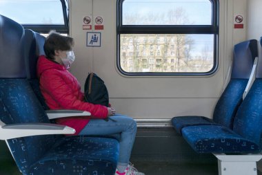 Tıbbi koruyucu maskeli genç bir kadın sırt çantasıyla tren koltuğunda oturuyor, diğer sandalyeler boş, etrafta kimse yok. Sosyal uzaklık, pandemia sırasında seyahat.
