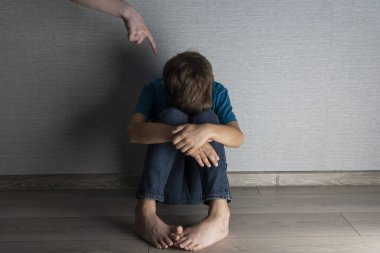 Scared boy seats on floor and hides his face in his knees, mother's hand is pointing with his finger above his head. Punishment, abuse and violence concept