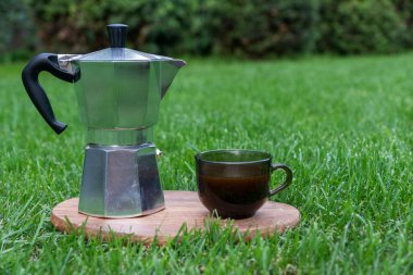 Demir gayzer kahve makinesi ve yeşil çimenlikteki ahşap tepside bir fincan kahve fotografı. Açık havada yaz kahvaltısı.