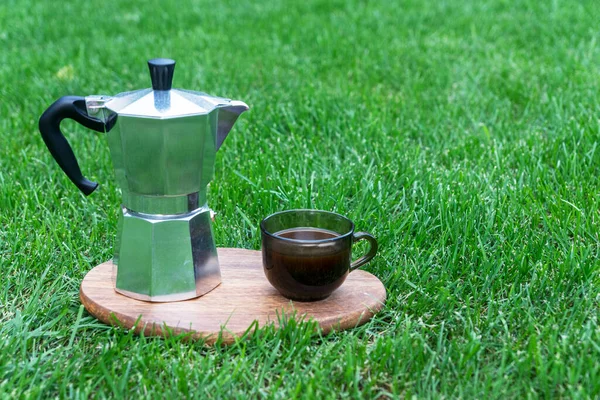 Morning outside coffee in summer. Iron geyser coffeemaker and cup of coffee on green grass, copy space.